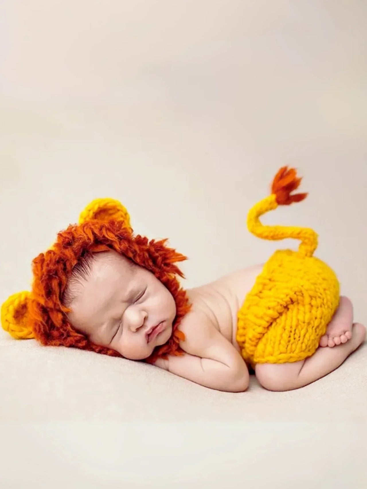 Newborn baby wearing a lion costume with a red mane and yellow body on a beige background