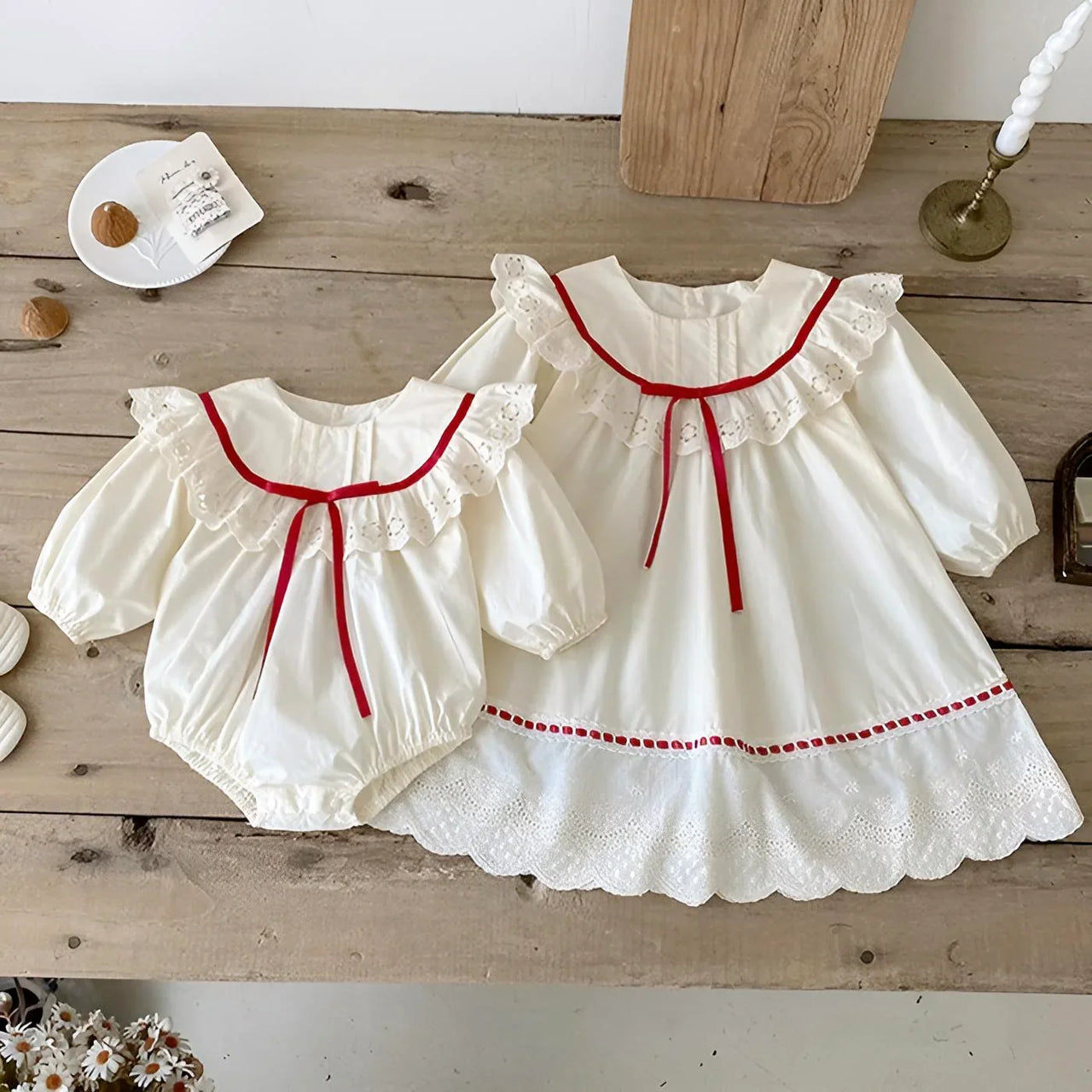Two white baby dresses with red trim on a wooden surface.