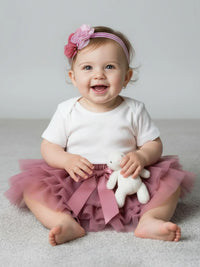 Thumbnail for Baby wearing a white onesie with a pink tutu and headband, holding a teddy bear.