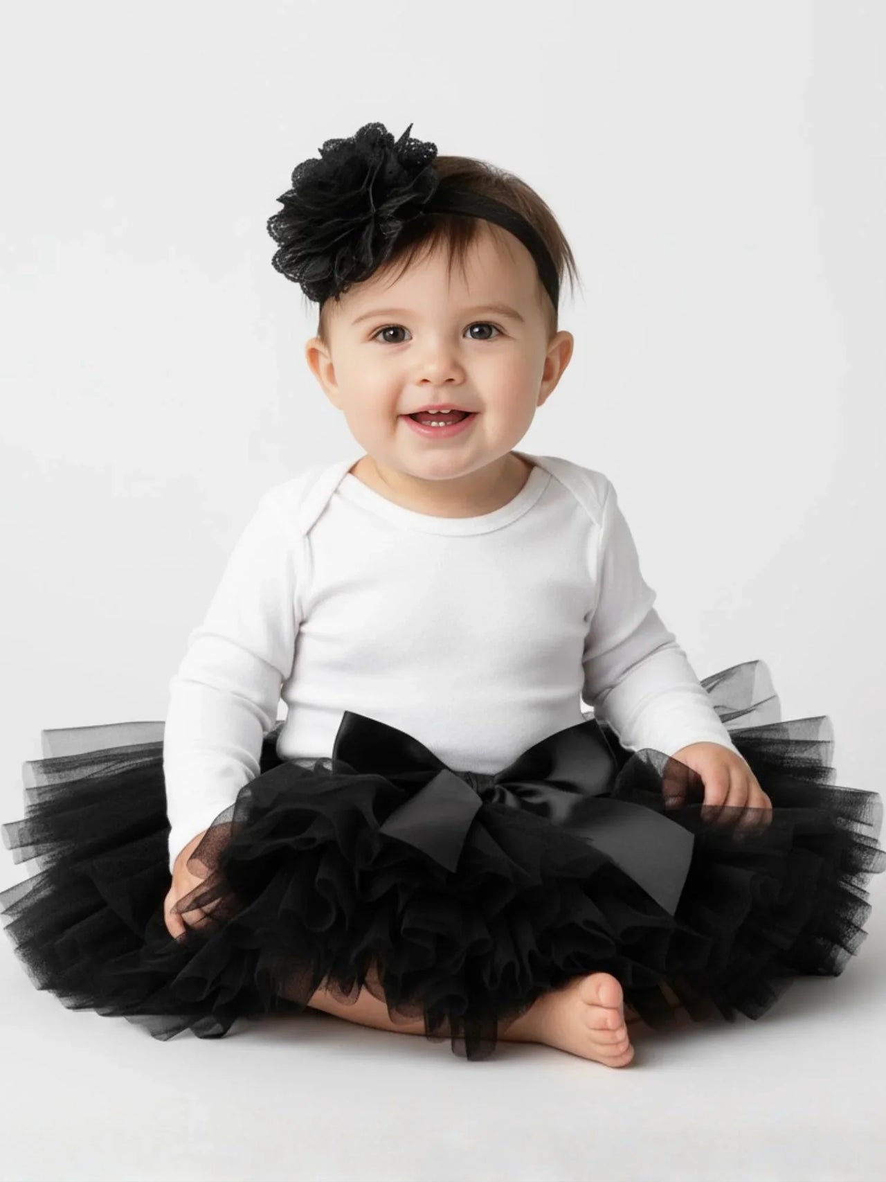 Baby wearing a white onesie with a black tutu and headband on a light gray background