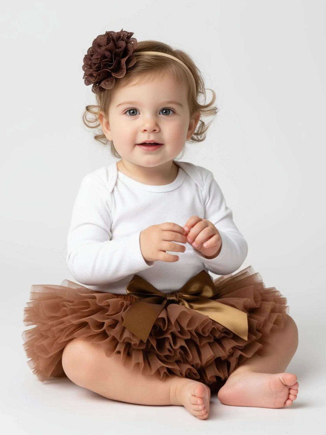 Baby wearing a white long-sleeve top and brown tutu skirt with a flower headband on a white background