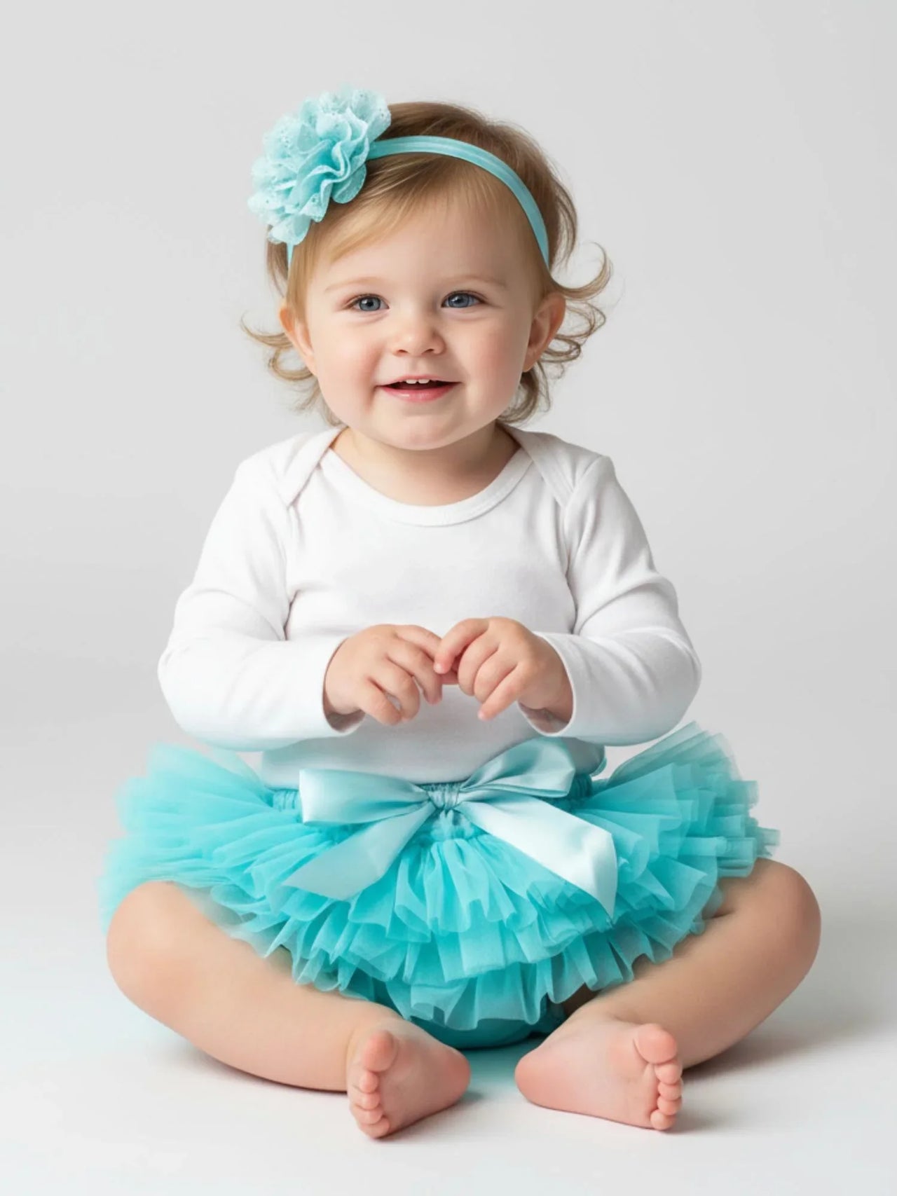 Baby wearing a white long-sleeve top and turquoise tutu with a matching headband on a light gray background