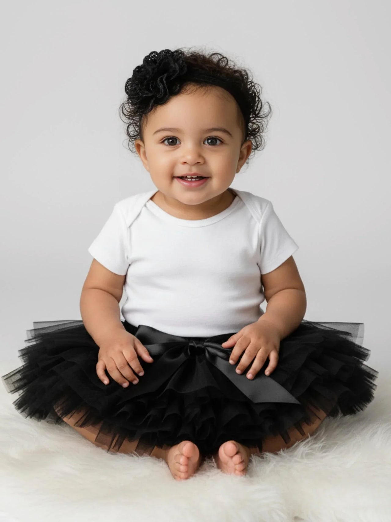 Baby wearing a white onesie with a black tutu sitting on a white surface.