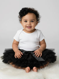 Thumbnail for Baby wearing a white onesie with a black tutu sitting on a white surface.