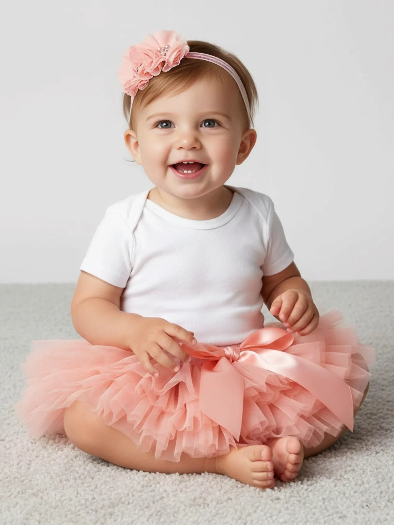 Baby wearing a white onesie with a pink tutu and headband on a light background