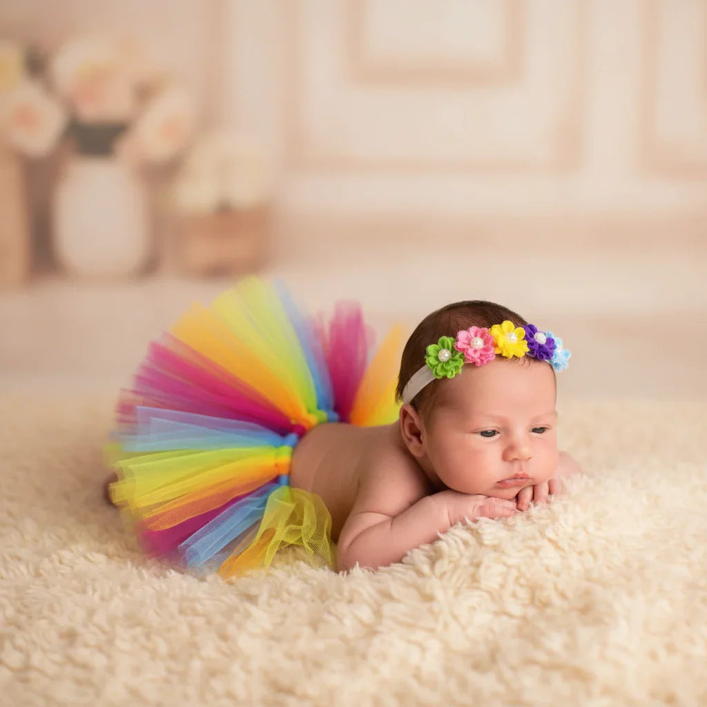 Newborn baby wearing a colorful rainbow tutu and floral headband on a soft surface.