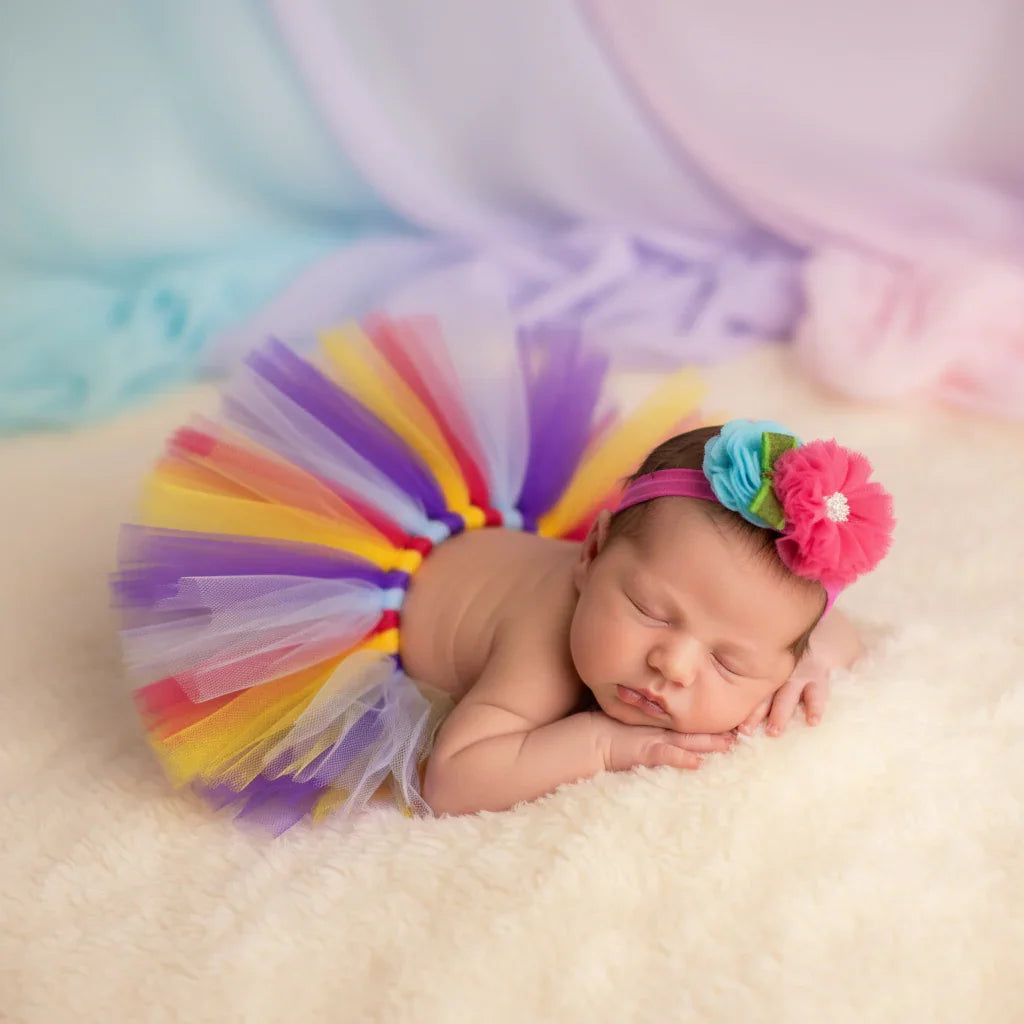 Newborn baby wearing a colorful tutu and headband on a soft surface with a pastel background