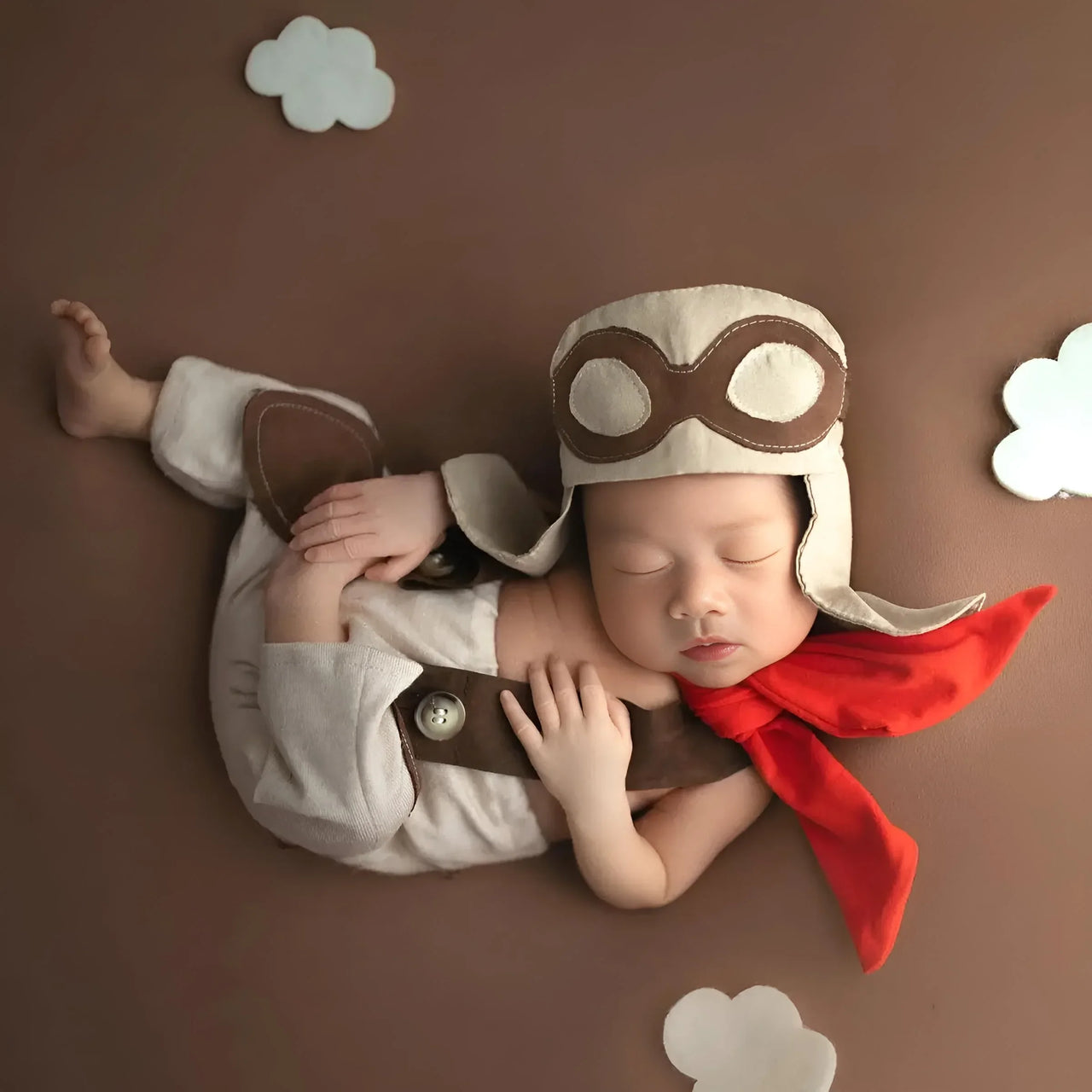 Newborn baby dressed as an aviator with a helmet and scarf on a brown background with cloud decorations.