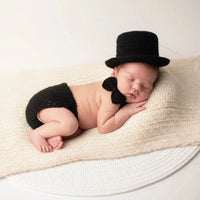 Thumbnail for Newborn baby wearing a black knitted outfit with a top hat and bow tie on a beige blanket.