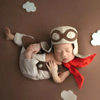 Thumbnail for Newborn baby dressed as an aviator with a helmet and scarf on a brown background with cloud decorations.