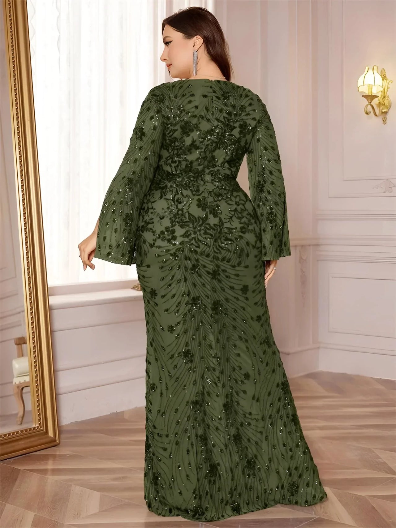 Plus size woman in an olive green evening gown standing next to a mirror in an elegant room.