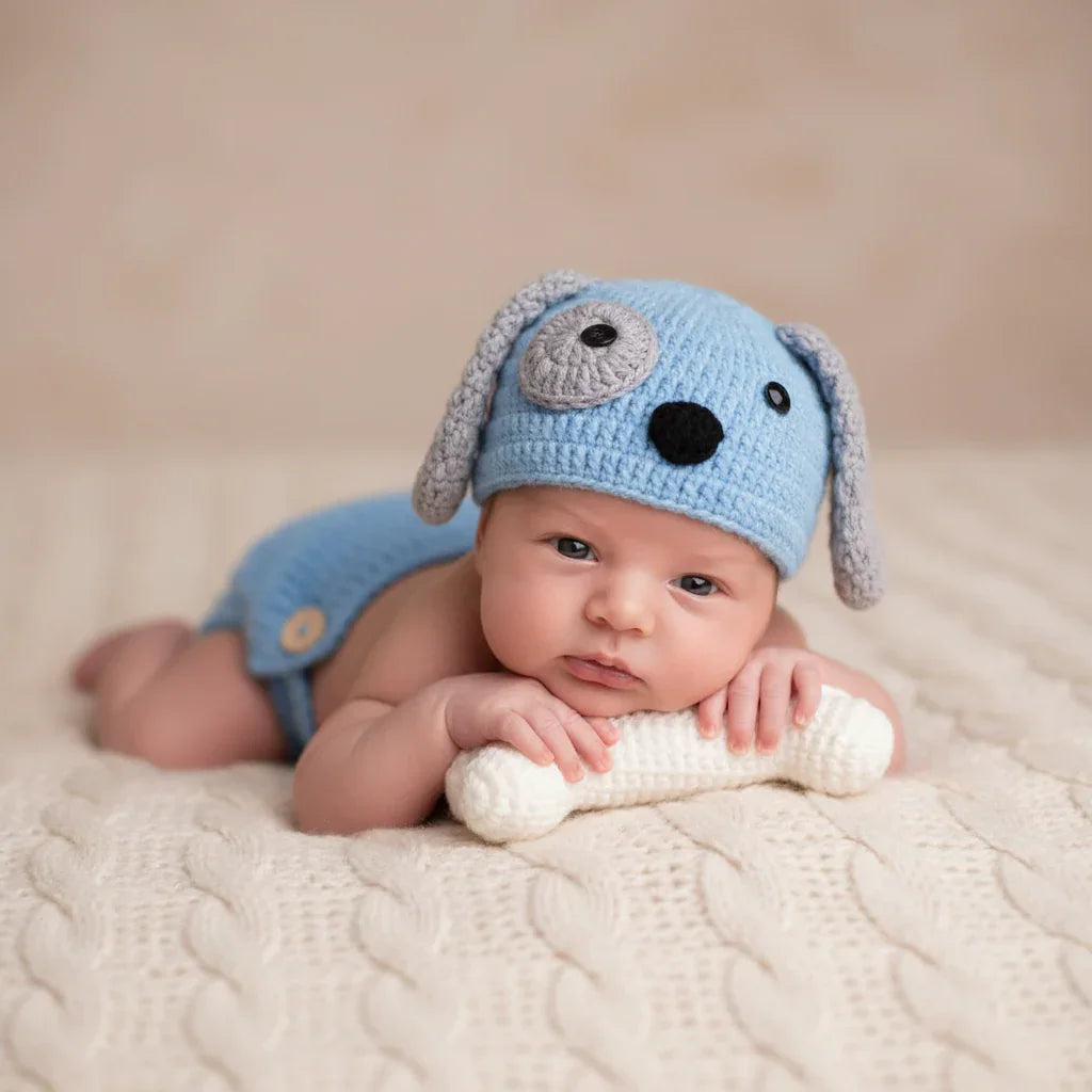 Newborn baby wearing a blue knitted dog hat and holding a white knitted toy on a beige blanket.
