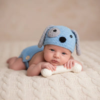 Thumbnail for Newborn baby wearing a blue knitted dog hat and holding a white knitted toy on a beige blanket.