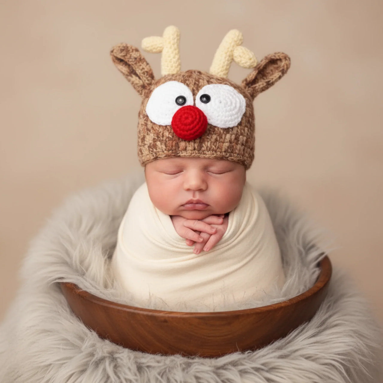 Newborn baby wearing a reindeer hat with antlers, white face, black eyes, and a red nose, wrapped in a white blanket in a wooden bowl.