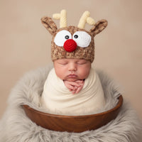 Thumbnail for Newborn baby wearing a reindeer hat with antlers, white face, black eyes, and a red nose, wrapped in a white blanket in a wooden bowl.