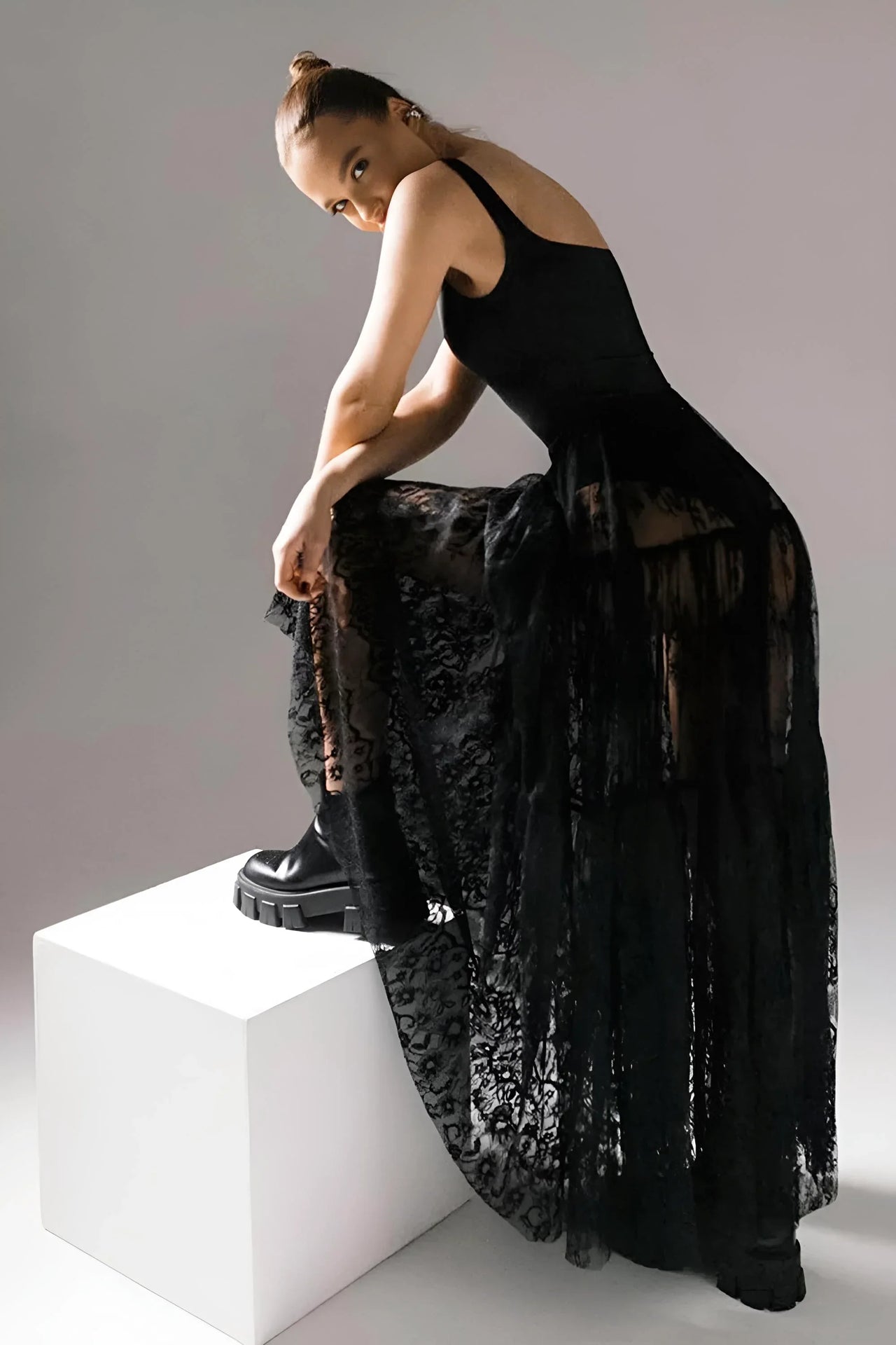 Woman in a black lace maxi skirt leaning on a white cube against a gray background