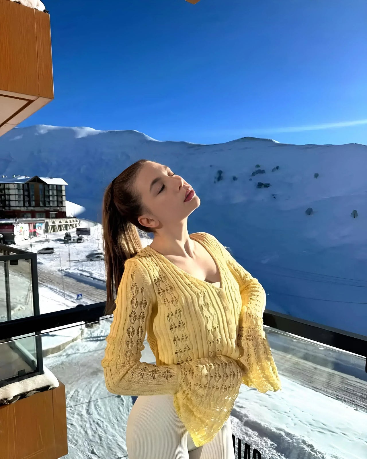 Woman in a yellow cardigan standing on a balcony with a snowy mountain landscape in the background