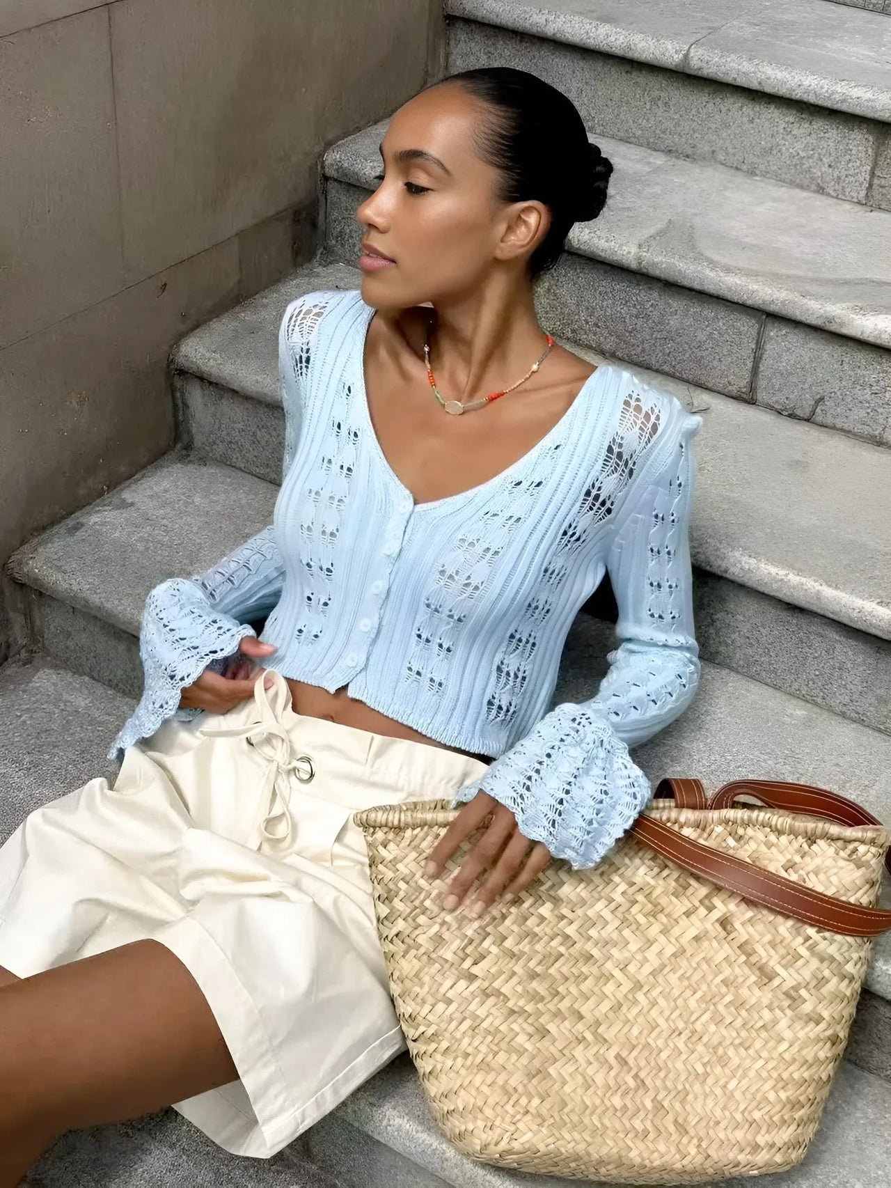 Woman sitting on steps wearing a light blue cardigan and white pants, holding a straw bag.
