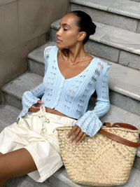 Thumbnail for Woman sitting on steps wearing a light blue cardigan and white pants, holding a straw bag.