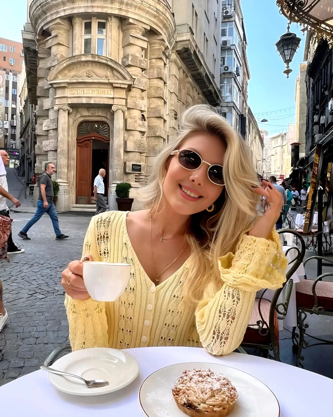 Woman in a yellow sweater sitting at an outdoor cafe table with a cup and pastry, smiling.