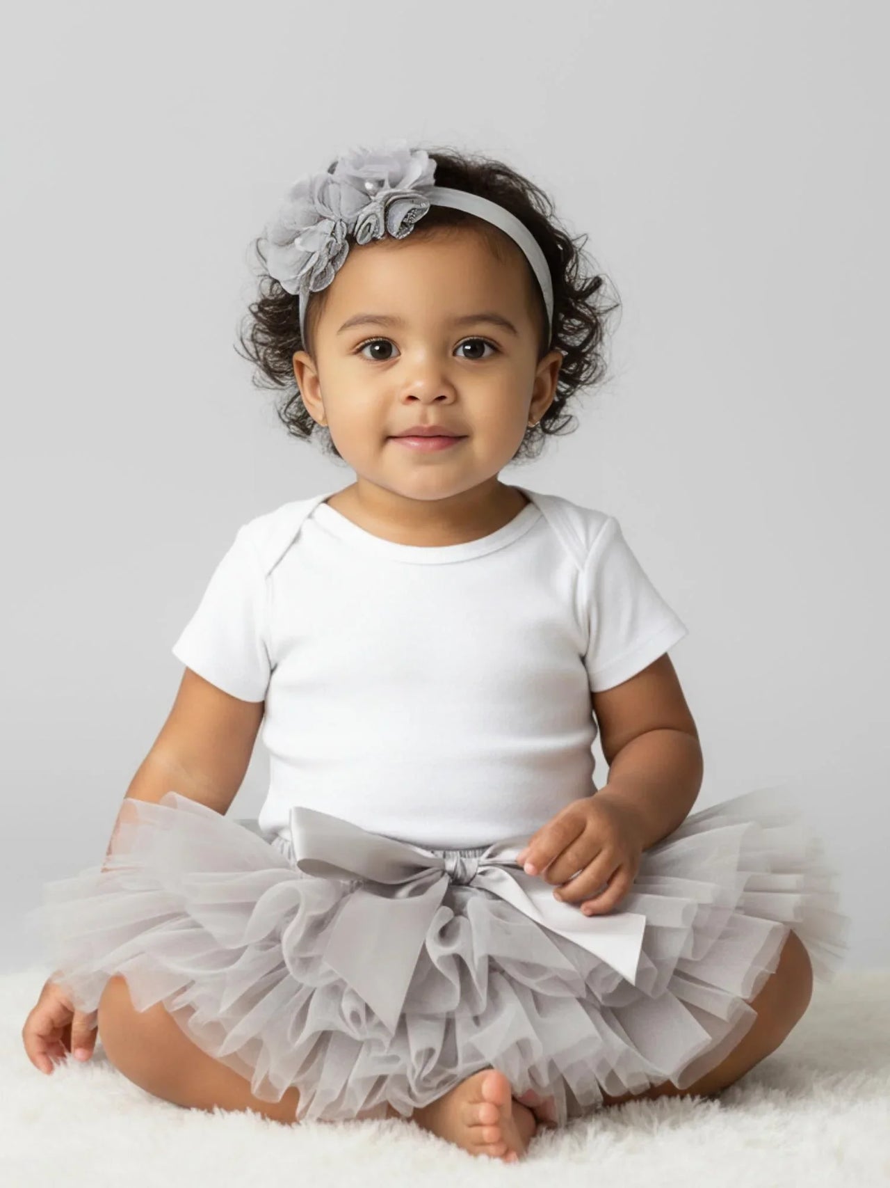 Baby wearing a white onesie with a gray tutu and headband on a light gray background