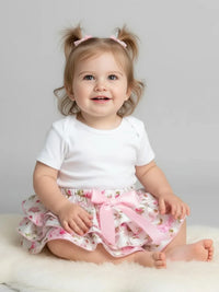Thumbnail for Child wearing a white shirt and floral bloomer with a pink bow, sitting on a white surface against a gray background.