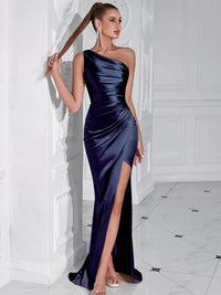 Thumbnail for Woman wearing a dark blue one-shoulder draped maxi dress with a high slit standing against a white wall.