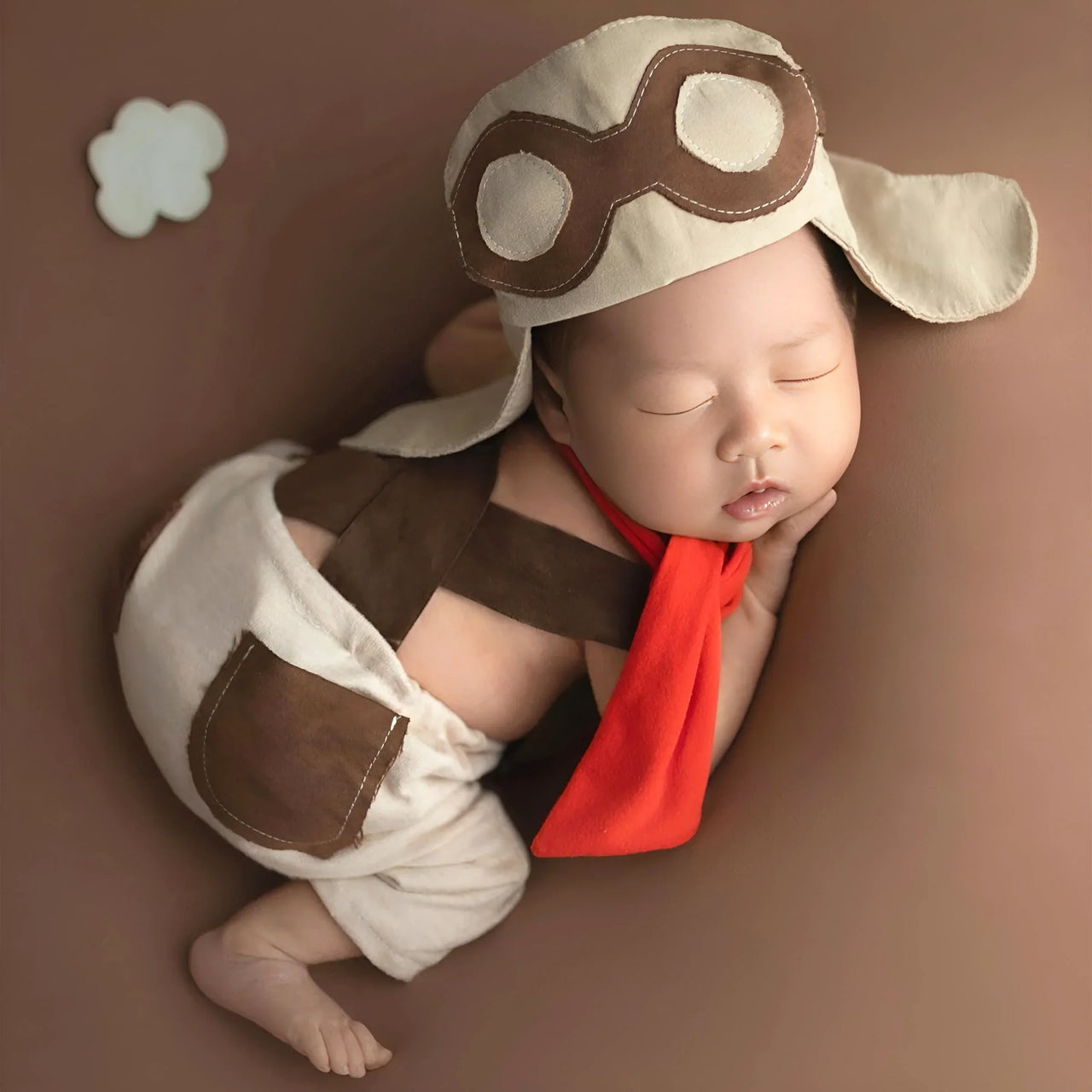 Newborn baby dressed in a superhero costume with a brown and white outfit and red cape on a brown background.