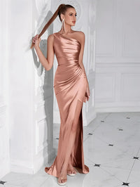 Thumbnail for Woman in a rose gold one shoulder draped maxi dress with a high slit standing against a white wall.