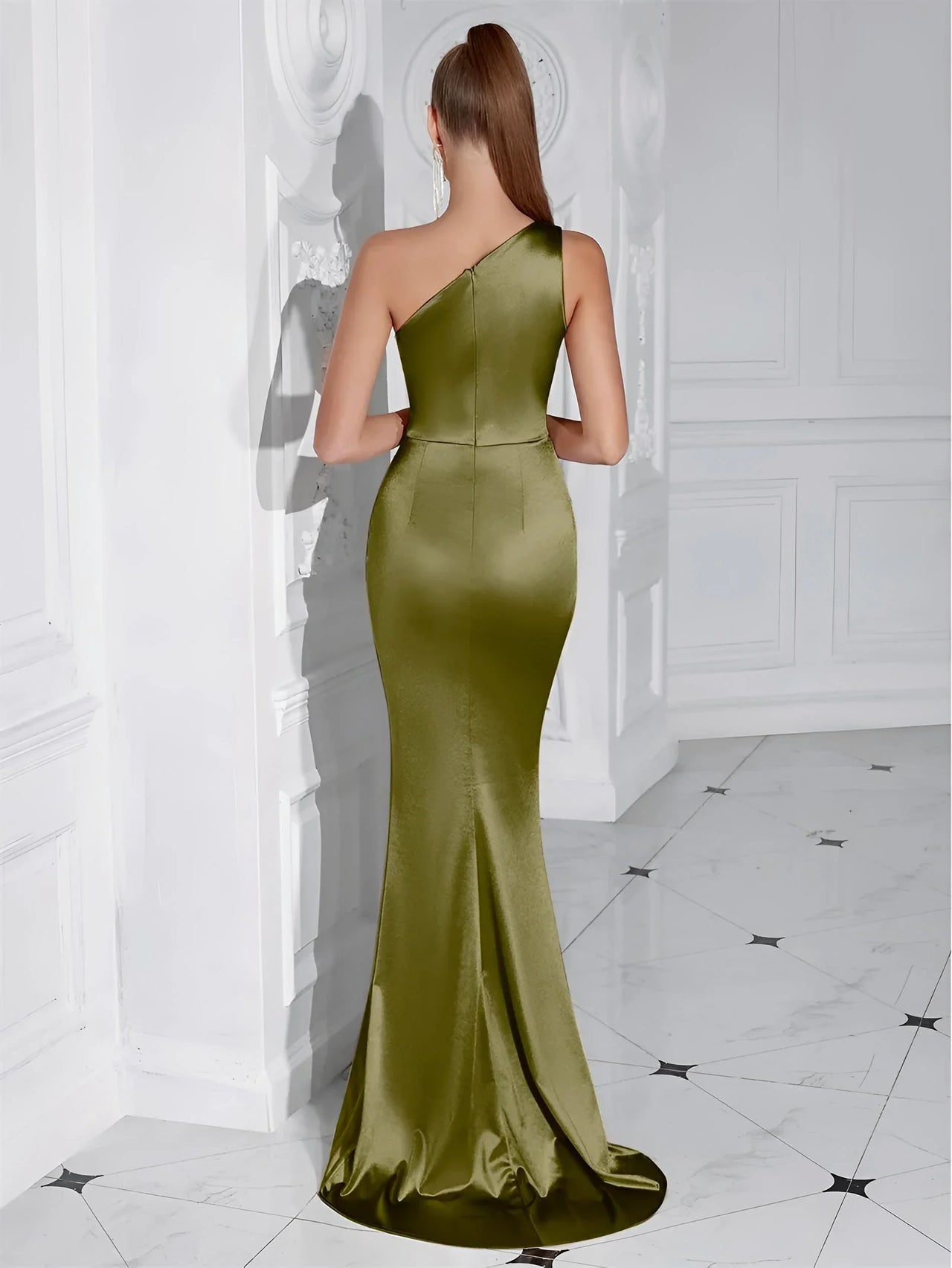 Back view of a woman wearing an olive green one-shoulder draped maxi dress in a white-walled room.