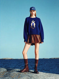 Thumbnail for Person wearing a navy teddy bear sweater, brown skirt, and boots standing on rocks with a blue sky and water background.