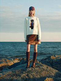 Thumbnail for Person standing on a rocky beach wearing a white teddy bear sweater with a design, brown skirt, and brown boots.