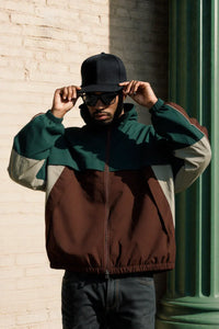 Thumbnail for Person wearing a green, brown, and beige jacket with a cap and sunglasses against a brick wall.