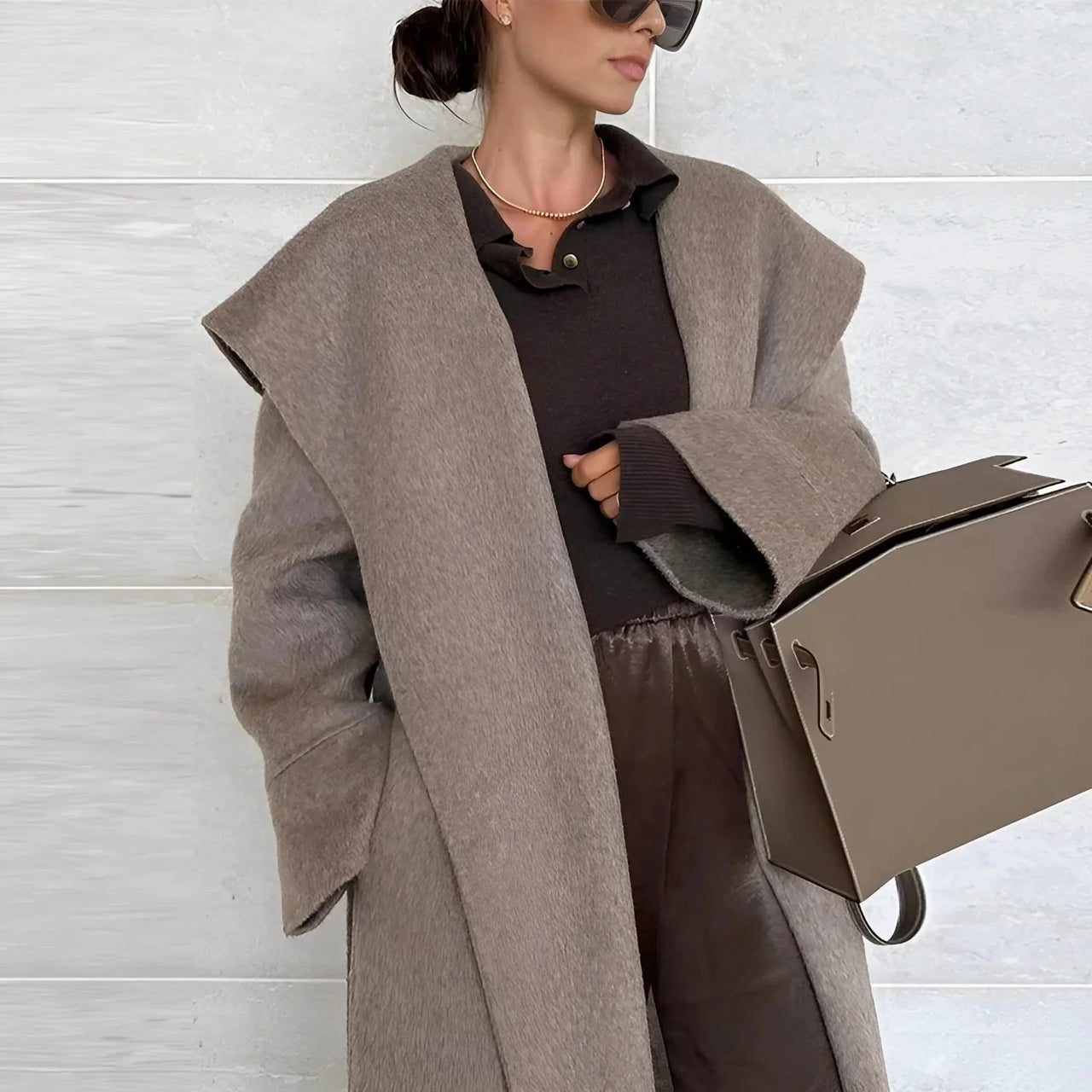 Woman wearing a long camel waterfall coat with belt and shawl lapel holding a beige purse on a white background